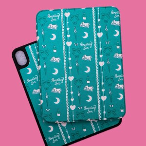May include: Teal tablet case with a repeating pattern of white hearts, bows, moons, books, and the text "Romantasy Lover." The case is open, revealing the interior and a black border.