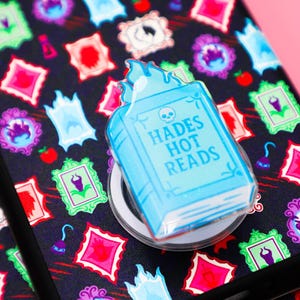 May include: A phone grip featuring a light blue book design with the text "HADES HOT READS" and a skull. The grip is on a phone case with a black background and colorful framed illustrations.