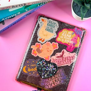 May include: A pink and clear glitter phone case with various colorful stickers. The stickers feature phrases like "Silly Goose on the Loose", "Let Me Read in Peace or I'll Scream", "Dreamer Nighttime Reader", and "Romantasy Girlie".