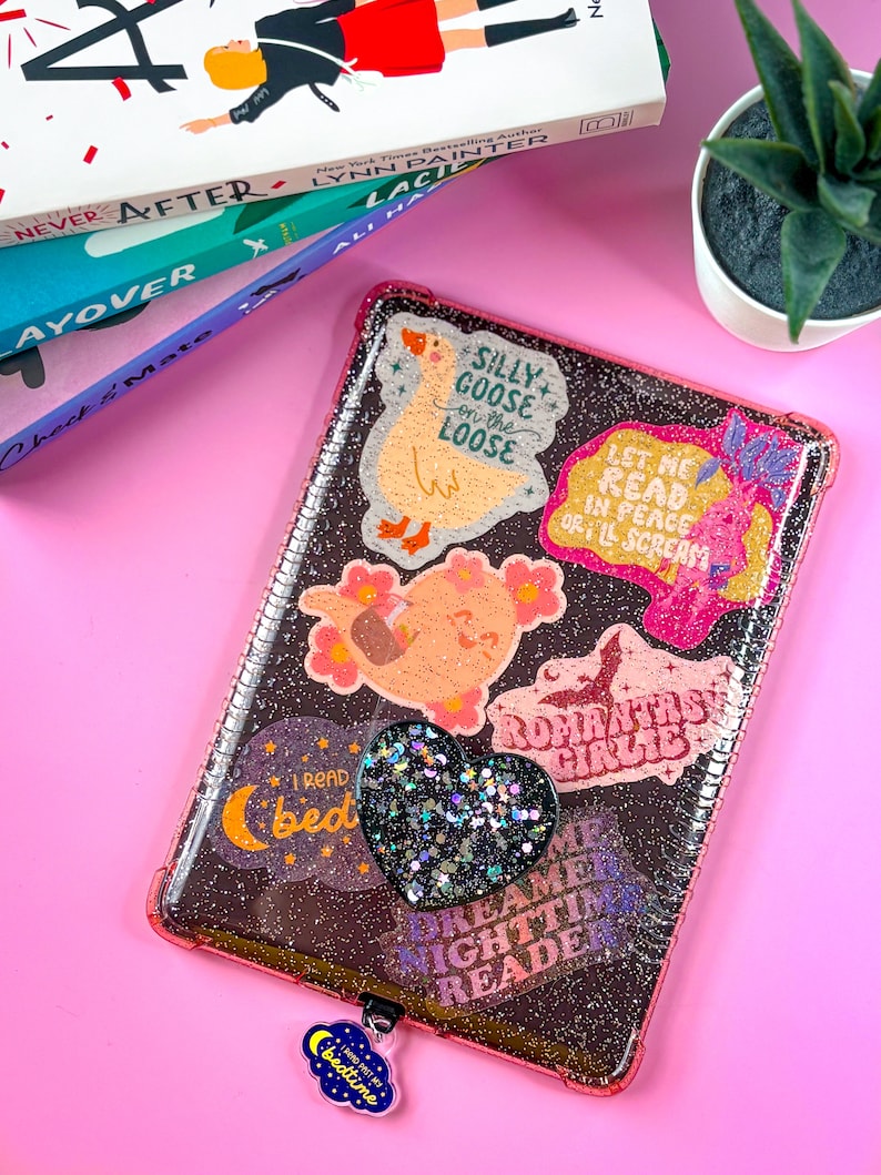 May include: A pink and clear glitter phone case with various colorful stickers. The stickers feature phrases like "Silly Goose on the Loose", "Let Me Read in Peace or I'll Scream", "Dreamer Nighttime Reader", and "Romantasy Girlie".