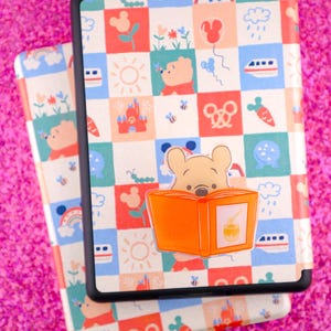 May include: Two tablet cases with a colorful Winnie the Pooh design. The cases feature a repeating pattern of squares with illustrations of Pooh, flowers, rainbows, and other Disney-themed images. A Pooh bear sticker is on the top case.