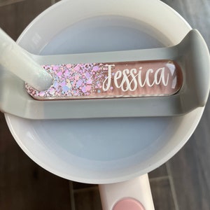 K&ouml;nnte beinhalten: Eine wei&szlig;e und graue Plastiktasse mit einem rosa Griff. Die Tasse hat einen transparenten Plastikeinsatz mit einem pink-wei&szlig;en Glitzerdesign und den Namen "Jessica" in wei&szlig;.
