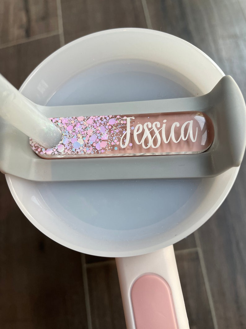 K&ouml;nnte beinhalten: Eine wei&szlig;e und graue Plastiktasse mit einem rosa Griff. Die Tasse hat einen transparenten Plastikeinsatz mit einem pink-wei&szlig;en Glitzerdesign und den Namen "Jessica" in wei&szlig;.