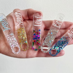 K&ouml;nnte beinhalten: F&uuml;nf durchsichtige Acryl-Flaschen&ouml;ffner mit eingearbeiteten Glitzer. Jeder Flaschen&ouml;ffner hat einen anderen Glitzerfarbton und einen darauf geschriebenen Namen:  "Jessie", "Sarah", "Liv", "Britt" und "Aileen".