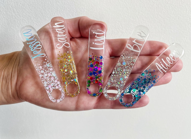 K&ouml;nnte beinhalten: F&uuml;nf durchsichtige Acryl-Flaschen&ouml;ffner mit eingearbeiteten Glitzer. Jeder Flaschen&ouml;ffner hat einen anderen Glitzerfarbton und einen darauf geschriebenen Namen:  "Jessie", "Sarah", "Liv", "Britt" und "Aileen".