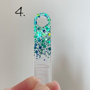 K&ouml;nnte beinhalten: Ein durchsichtiger Acryl-Schl&uuml;sselanh&auml;nger mit einem glitzernden Design in Gr&uuml;n und Blau. Der Schl&uuml;sselanh&auml;nger hat oben ein Loch zum Anbringen an einem Schl&uuml;sselring.