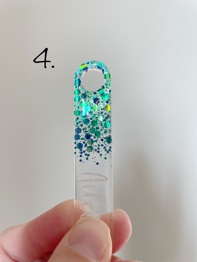 K&ouml;nnte beinhalten: Ein durchsichtiger Acryl-Schl&uuml;sselanh&auml;nger mit einem glitzernden Design in Gr&uuml;n und Blau. Der Schl&uuml;sselanh&auml;nger hat oben ein Loch zum Anbringen an einem Schl&uuml;sselring.