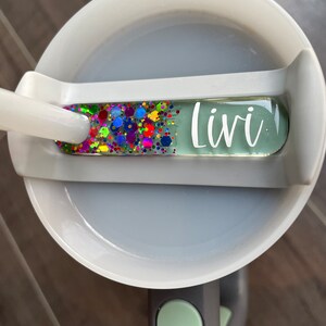 K&ouml;nnte beinhalten: Eine wei&szlig;e Keramiktasse mit einem gr&uuml;nen Deckel und einem durchsichtigen, bunten, glitzernden Namensschild mit der Aufschrift "Livi".