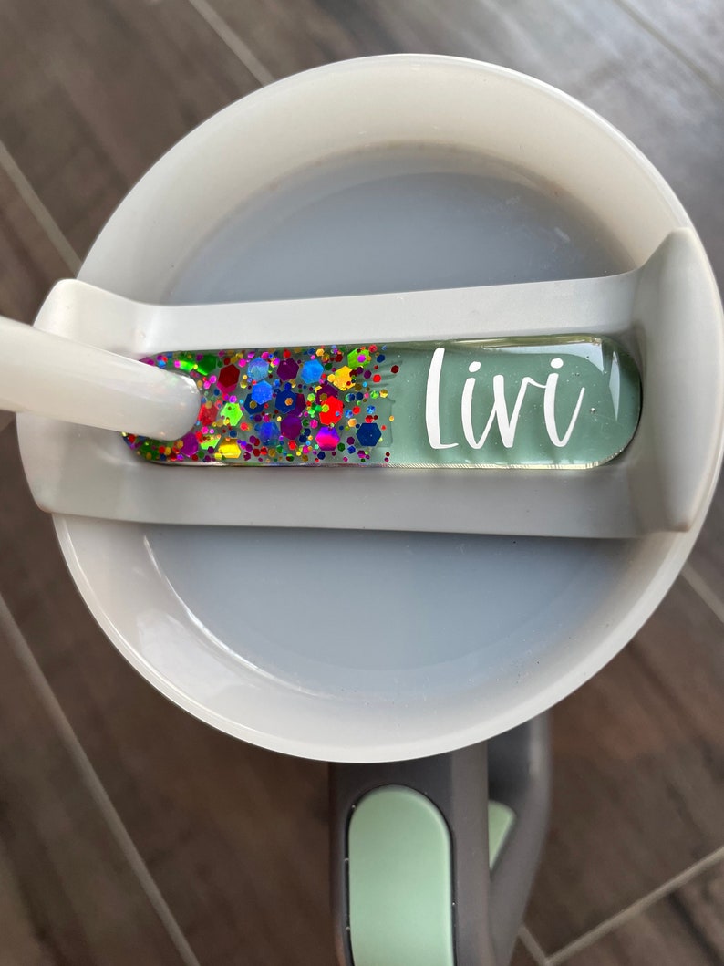 K&ouml;nnte beinhalten: Eine wei&szlig;e Keramiktasse mit einem gr&uuml;nen Deckel und einem durchsichtigen, bunten, glitzernden Namensschild mit der Aufschrift "Livi".