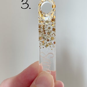 K&ouml;nnte beinhalten: Ein transparenter Glas-Anh&auml;nger mit goldenem Glitzer-Design und dem Wort "Smile" darauf. Der Anh&auml;nger hat oben eine Schlaufe zum Aufh&auml;ngen.