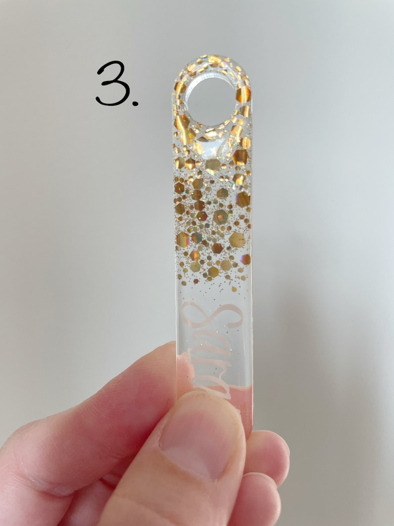 K&ouml;nnte beinhalten: Ein transparenter Glas-Anh&auml;nger mit goldenem Glitzer-Design und dem Wort "Smile" darauf. Der Anh&auml;nger hat oben eine Schlaufe zum Aufh&auml;ngen.