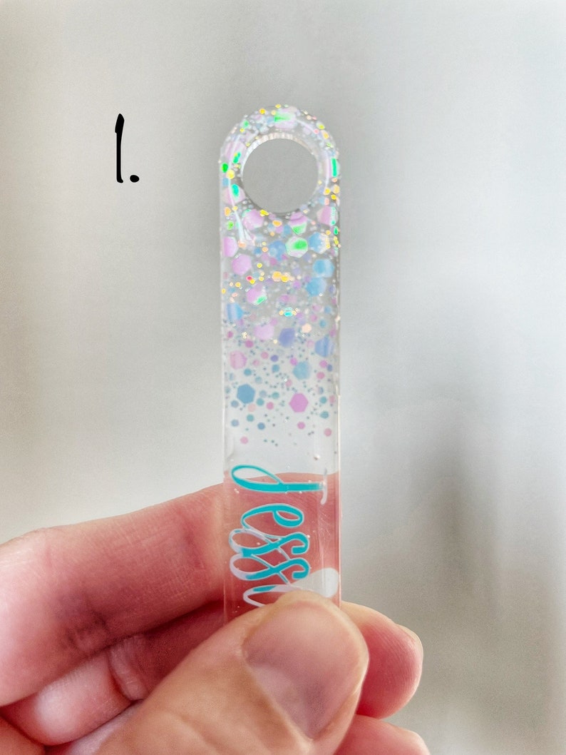 K&ouml;nnte beinhalten: Ein transparenter, rechteckiger Schl&uuml;sselanh&auml;nger mit einem Loch oben. Der Schl&uuml;sselanh&auml;nger ist mit schimmerndem Glitzer und bunten Konfetti gef&uuml;llt. Der Name "Jess" ist in t&uuml;rkisfarbener Schrift auf dem Schl&uuml;sselanh&auml;nger geschrieben.