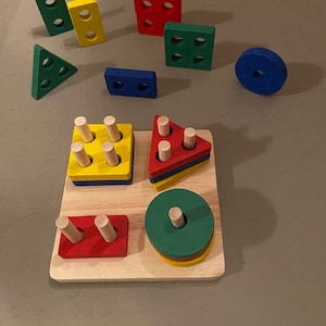 May include: Wooden shape sorter toy with a variety of colorful shapes, including a circle, square, triangle, and rectangle. The toy has a base with pegs for the shapes to be placed on.