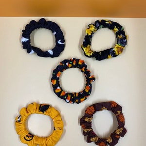 May include: Five fabric scrunchies in various fall-themed prints. The scrunchies are black with white ghosts, black with yellow sunflowers, black with candy corn, yellow with black jack-o-lanterns, and brown with orange and yellow leaves.