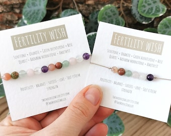 May include: Two cards with the text "Fertility Wish" and a list of gemstones. The cards are next to each other and have a string bracelet with gemstones on them. The bracelet has sunstone, unakite, green aventurine, rose quartz, rainbow moonstone, and amethyst.