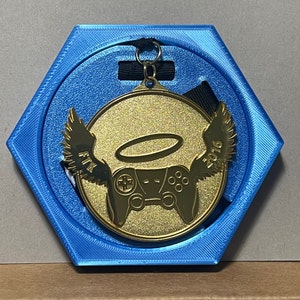 May include: A gold medal featuring a controller design, wings, and a halo, set in a blue hexagonal case. The medal displays the year 2016 and is attached to a black ribbon.