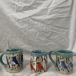May include: Three ceramic mugs with bird designs. The mugs have a light beige base with blue and red bird illustrations. The rims and handles are painted in a teal colour. The mugs are numbered 1, 2, and 3.