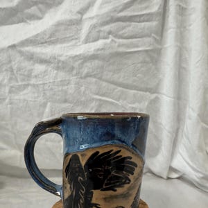 Può includere: Tazza in ceramica fatta a mano con un disegno di corvo. La tazza presenta una glassa blu scuro sulla parte superiore e una base marrone con un'illustrazione di corvo nero. Il manico è anch'esso blu scuro. Il corvo è raffigurato con dettagli intricati.