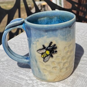May include: Handmade ceramic mug with a blue and cream glaze. The mug features a sculpted bee and honeycomb texture. The handle is curved and has a leaf-like detail. A unique, handcrafted piece for coffee or tea.