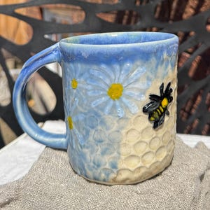 May include: A blue and white ceramic mug with a bee and daisy design. The mug has a textured surface that resembles a honeycomb.