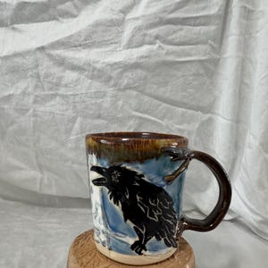 Könnte beinhalten: Handgefertigte Keramik-Tasse mit einem schwarzen Raben-Design vor blau-weißem Hintergrund. Die Tasse hat einen braunen Henkel und Rand und steht auf einem mehrschichtigen, strukturierten Sockel. Ein einzigartiges, handgefertigtes Stück für Kaffee oder Tee.