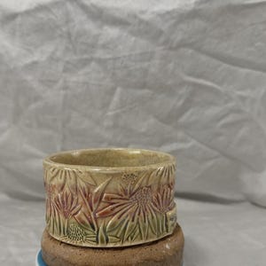 Peut inclure: Un bol en céramique beige avec un motif floral. Le bol présente des fleurs en relief dans des tons de rouge, vert et beige. Le bol cylindrique repose sur une base marron. L'ensemble dégage une impression rustique et artisanale.