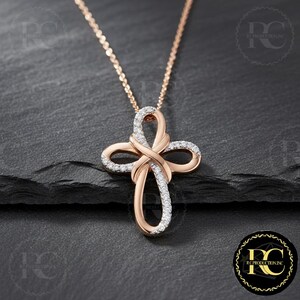 May include: Rose gold cross pendant necklace with a delicate chain. The cross features an intertwined design, embellished with sparkling white gemstones. The pendant is displayed on a dark, textured surface.