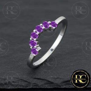 Könnte beinhalten: Ein silberner Ring mit sechs runden, leuchtend violetten Edelsteinen, die in einer gebogenen Reihe angeordnet sind. Der Ring hat eine polierte, reflektierende Oberfläche, und die Edelsteine sind gleichmäßig verteilt. Der Ring liegt auf einer dunklen, strukturierten Oberfläche.