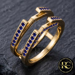 May include: A gold ring guard featuring two parallel bands connected by a bridge, each set with a row of small, round, vibrant blue gemstones. The design creates an open space intended to frame another ring, showcasing a polished metallic finish.