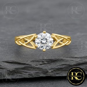 May include: A gold engagement ring with a large, round, clear gemstone. The ring has a Celtic knot design on the band. The ring is set against a dark grey background.