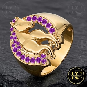 Puede incluir: Un anillo dorado con un diseño de caballo, rodeado por una media luna de piedras preciosas moradas. El anillo tiene una banda ancha y un acabado pulido. El caballo está representado en posición reclinada, con un diseño detallado.