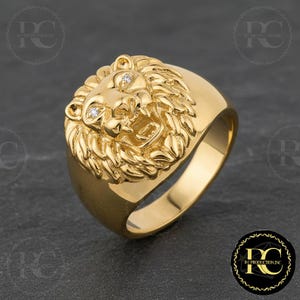 Può includere: Un anello dorato con un'elaborata testa di leone, con due piccoli accenti scintillanti. L'anello ha una fascia larga e una finitura lucida, che mette in risalto la criniera e i lineamenti del leone.