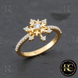 May include: A gold-colored ring featuring a snowflake design. The center of the snowflake has a round, clear gemstone, with smaller gemstones on the snowflake's points and along the ring band. The ring is set against a dark background.