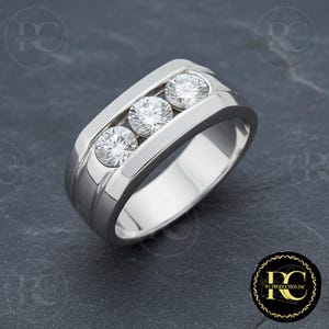 May include: A silver ring with three round, clear gemstones set in a channel. The ring has a polished finish and a wide band with subtle linear detailing. The gemstones are evenly spaced and catch the light.
