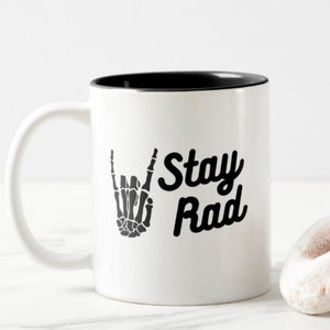 May include: White ceramic coffee mug with a black rim. The mug has a black and white graphic of a skeleton hand making a rock and roll hand gesture. The text "Stay Rad" is printed in black on the mug.