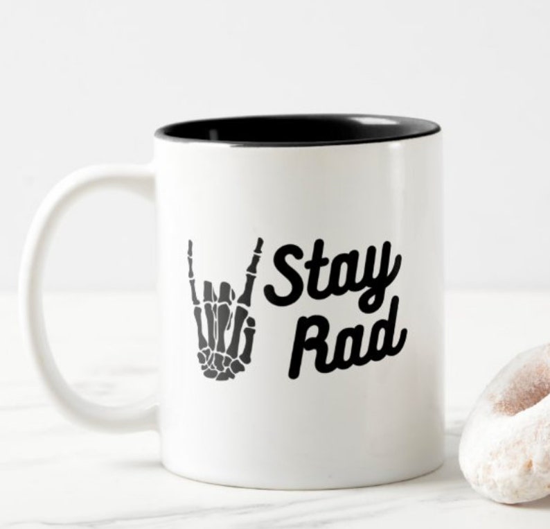May include: White ceramic coffee mug with a black rim. The mug has a black and white graphic of a skeleton hand making a rock and roll hand gesture. The text "Stay Rad" is printed in black on the mug.