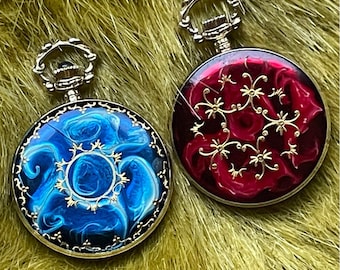 May include: Two pocket watches with ornate designs. One watch features a blue, swirling pattern, while the other has a red background with gold floral accents. Both have silver-colored metal casings and loops.
