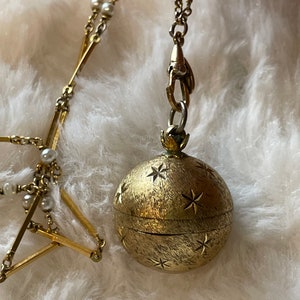 Puede incluir: Un collar dorado con un colgante redondo adornado con recortes de estrellas y una superficie texturizada. El collar incluye una delicada cadena con pequeñas perlas y barras geométricas doradas. El colgante está sujeto a una cadena con un cierre decorativo.