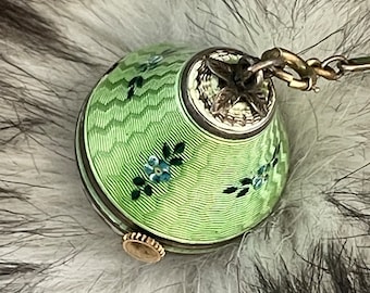 May include: A small, antique, green enameled compact with a floral design. The compact has a silver-colored metal frame and a gold-colored clasp. A delicate chain is attached to the top. The compact is resting on a fluffy, gray and white surface.