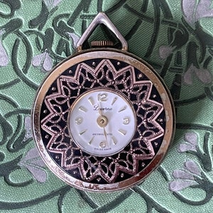 Puede incluir: Un reloj de bolsillo Lucerne vintage con esfera blanca y diseño de filigrana negra. El reloj tiene una caja de metal plateado y un pequeño lazo en la parte superior para una cadena. La esfera muestra la hora con números y agujas negras.