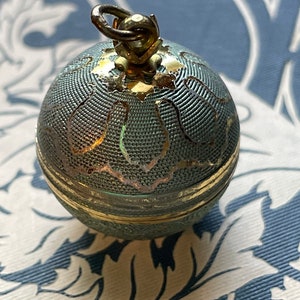 Puede incluir: Una pequeña y redonda caja de baratijas antigua con tapa con bisagras. La caja es de color verde azulado con un diseño de filigrana dorada y una banda dorada alrededor del centro. Tiene un pequeño lazo en la parte superior para colgar.