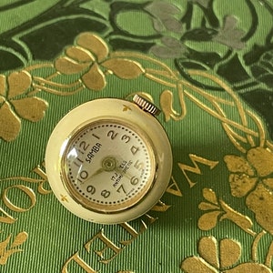 Puede incluir: Un pequeño reloj vintage redondo con esfera color crema y bisel dorado. La esfera del reloj muestra la palabra "SAMBA" y números. El reloj está sobre un fondo verde con detalles florales dorados.