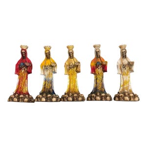 May include: Five colorful statues of a robed skeleton figure wearing a crown, each standing on a base of skulls. The figures are in red, yellow, gold, orange, and white, with glitter accents on their robes.