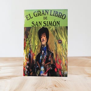 May include: A book titled "EL GRAN LIBRO DE SAN SIMÓN" with a green and black cover. The cover features a man in a black hat and suit, surrounded by various objects. The background is a mix of red and gold.