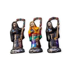May include: Three colorful Santa Muerte statues. Each figure wears a hooded robe and holds a staff topped with flowers. The central statue has a rainbow-colored robe, while the others are black. The statues have a skull face.