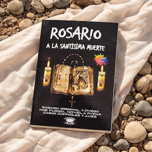 May include: A black book titled "ROSARIO A LA SANTISIMA MUERTE" with gold illustrations of candles and an open book. The book is resting on a white cloth, with rocks in the background.