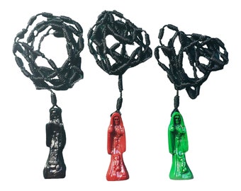 May include: Three beaded necklaces with pendants. Each necklace has a black, woven rope design. The pendants are in the shape of a robed figure, one black, one red, and one green. The pendants are suspended from the rope.