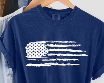 Puede incluir: Una camiseta azul marino con un diseño de bandera estadounidense blanca desgastada. La camiseta está doblada y colgada en una percha. El texto "TRUE NAVY" es visible en la parte inferior de la imagen.