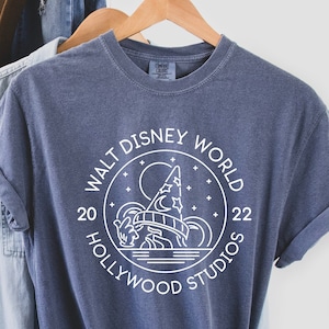 Puede incluir: Una camiseta azul marino con un gr&aacute;fico blanco del logotipo de Walt Disney World Hollywood Studios. El logotipo presenta una silueta de las ic&oacute;nicas colinas de Hollywood y un sombrero de mago. El texto "Walt Disney World" est&aacute; escrito en un c&iacute;rculo alrededor del gr&aacute;fico, con los a&ntilde;os "20" y "22" a cada lado.