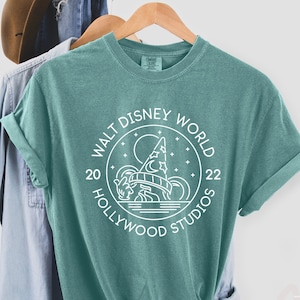 Puede incluir: Una camiseta azul turquesa con un dise&ntilde;o gr&aacute;fico blanco en forma de c&iacute;rculo con el texto "Walt Disney World 20 Hollywood Studios 22" y una ilustraci&oacute;n estilizada del Hollywood Tower Hotel.